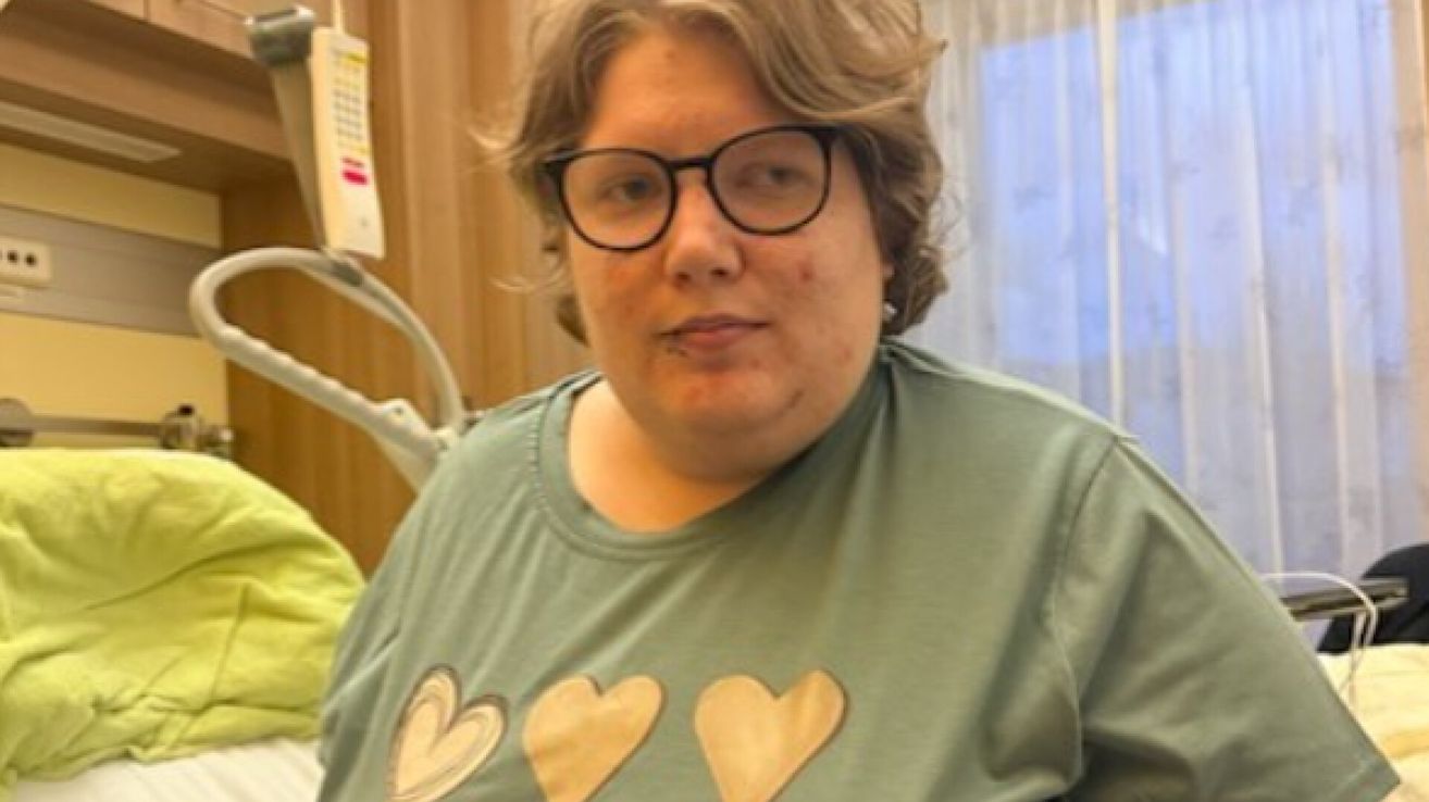Eine Frau mit kurzen Haaren und Brille trägt ein grünes T-Shirt mit Herzdesigns und sieht direkt in die Kamera. Sie befindet sich in einem Raum mit einer Holzwand und einem weißen Vorhang.