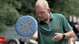 Ein älterer Mann in einem grünen Poloshirt hält ein blaues kreisförmiges Objekt, möglicherweise ein Frisbee, mit einem dekorativen Muster. Er schaut zur Seite und erklärt möglicherweise etwas.