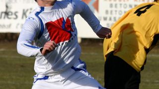 Ein Fußballspieler in einem weißen Trikot mit der Nummer 13 läuft auf einem Feld. Ein weiterer Spieler in einem gelben Trikot steht neben ihm. Dahinter ist ein weißes Banner mit Text.