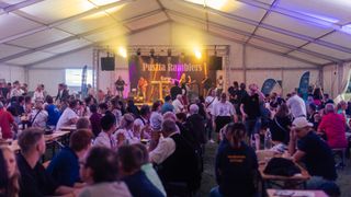 Eine Band spielt auf der Bühne in einem Zelt, mit gelber Beleuchtung. Ein Publikum von Menschen sitzt vor der Bühne, einige klatschen. Die Bühne hat ein Banner mit dem Text 'Puszta Ramblers'.