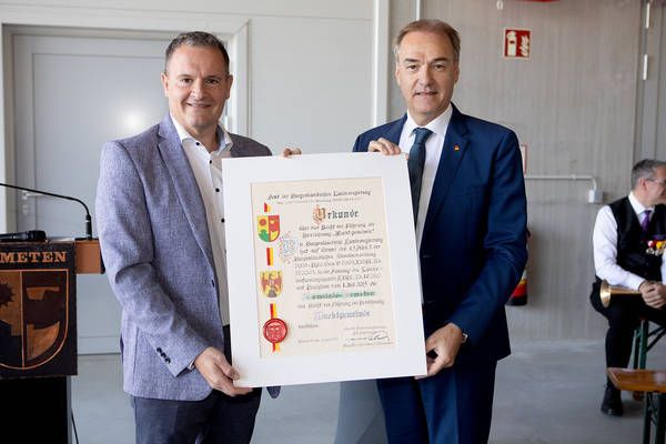 Zwei Männer in Anzügen halten ein gerahmtes Dokument, lächeln, mit einem Mikrofon und einem Podium im Hintergrund.