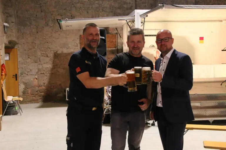 Drei Männer stehen zusammen, lächeln und halten Biergläser in der Hand. Sie befinden sich wahrscheinlich bei einer Bierveranstaltung. Der Mann auf der linken Seite trägt eine schwarze Uniform mit Logo. Der Mann in der Mitte trägt ein schwarzes T-Shirt. Der Mann auf der rechten Seite trägt einen Anzug.