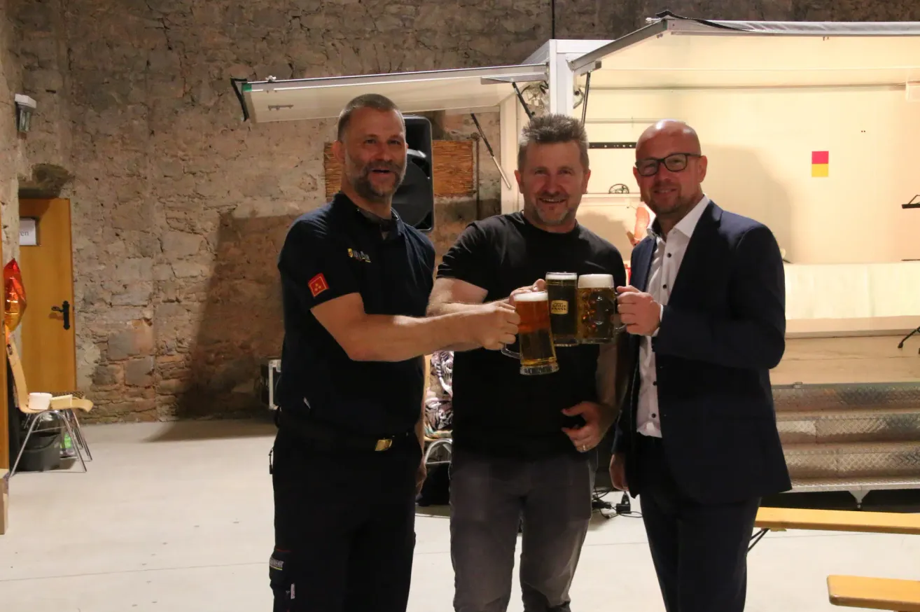 Drei Männer stehen zusammen, lächeln und halten Biergläser in der Hand. Sie befinden sich wahrscheinlich bei einer Bierveranstaltung. Der Mann auf der linken Seite trägt eine schwarze Uniform mit Logo. Der Mann in der Mitte trägt ein schwarzes T-Shirt. Der Mann auf der rechten Seite trägt einen Anzug.