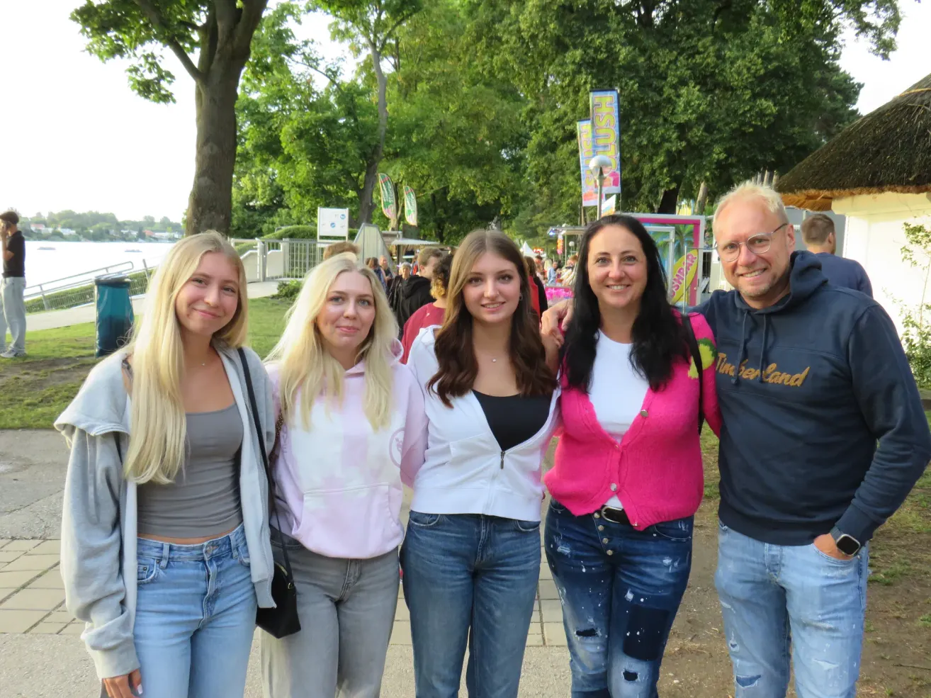Vier Frauen und ein Mann stehen mit lächelndem Gesicht im Park. Die Frau links hält eine Handtasche. Sie sind von Bäumen umgeben. In der Ferne gehen einige Leute. Es gibt einige Werbetafeln, die an den Pfosten montiert sind.