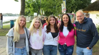 Vier Frauen und ein Mann stehen mit lächelndem Gesicht im Park. Die Frau links hält eine Handtasche. Sie sind von Bäumen umgeben. In der Ferne gehen einige Leute. Es gibt einige Werbetafeln, die an den Pfosten montiert sind.