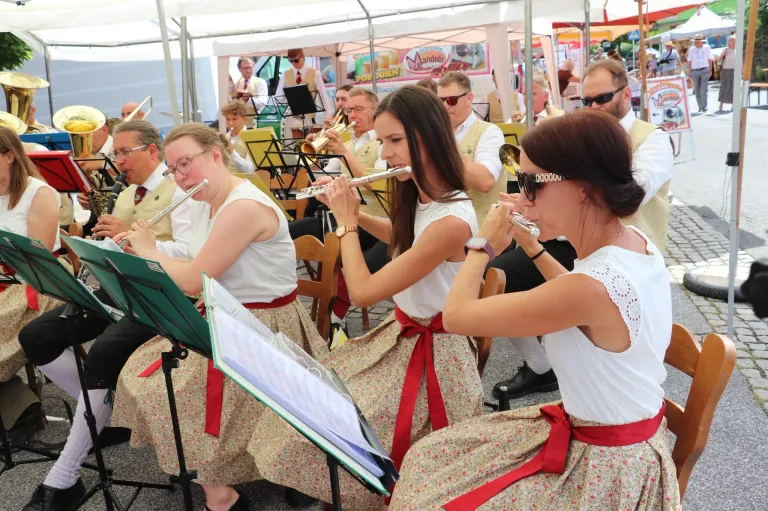 Eine Gruppe von Frauen in weißen Oberteilen und Blumenröcken mit roten Bändern spielt Blasinstrumente. Sie sitzen auf einer Bank mit Notenständern. Hinter ihnen spielen andere Musiker Instrumente. Dahinter steht ein Zelt mit einem Händler.