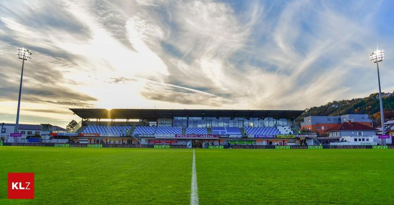 Ein leeres Fußballstadion mit blauen Sitzen und einem großen Spielfeld. Der Himmel ist bewölkt mit einem untergehenden Sonnenlicht sichtbar.
