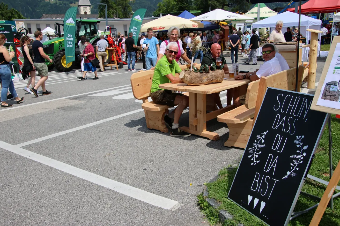 Mensch sitzt an Picknicktischen bei einer Outdoor-Veranstaltung. Ein Mann in einem grünen Shirt sitzt mit einem Glas Bier. Dahinter sind Zelte und ein Traktor. Ein Schild auf der rechten Seite sagt 'So DA DA BIS'.
