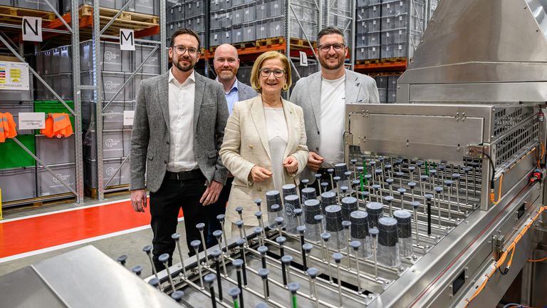 Fünf Personen, in formeller Kleidung gekleidet, stehen vor einer Maschine, die Behälter füllt. Sie lächeln für die Kamera. Dahinter befindet sich ein Lager mit Lagerboxen in den Regalen.