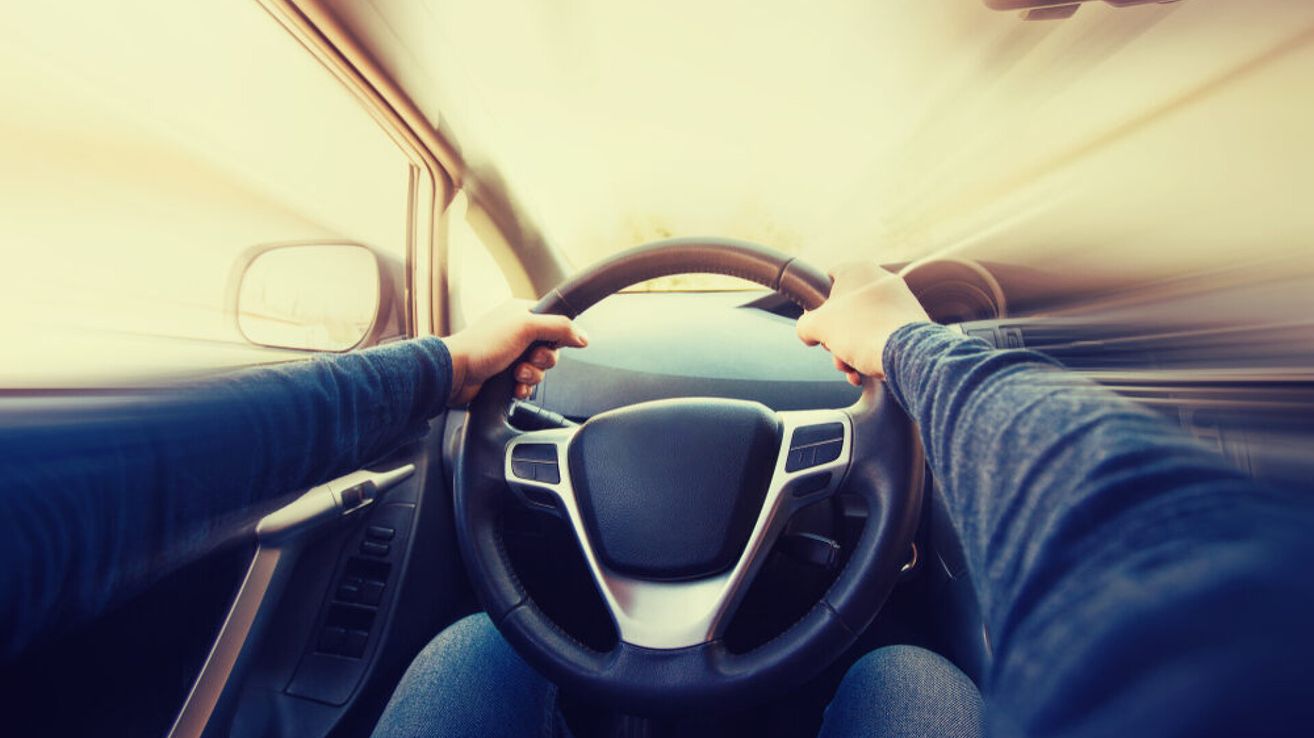 Eine Person fährt ein Auto mit beiden Händen am Lenkrad, der Armaturenbrett ist im Vordergrund sichtbar.