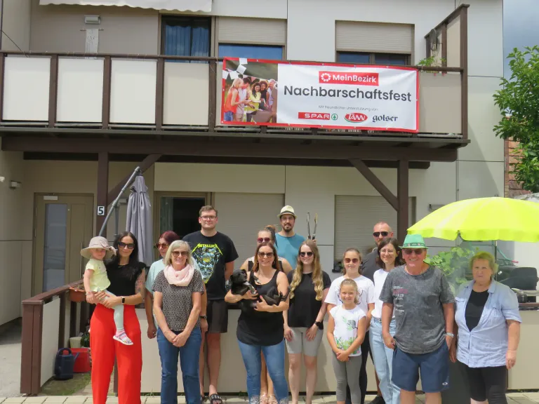 Eine Gruppe von Menschen und ein Hund posieren für ein Foto vor einem Gebäude mit einem Banner, das Nachbarschaftsfest liest.