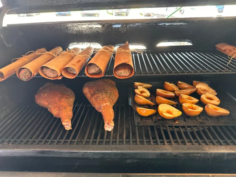 Ein Outdoor-Grill mit zwei gewürzten Fleischstücken und einer Schale mit Apfelscheiben, alles in Bananenblätter gewickelt und bereit zum Kochen.