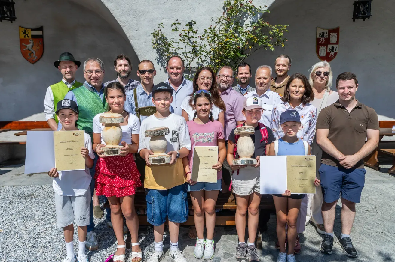 Eine Gruppe von Menschen steht vor einem Gebäude mit einer Pflanze, lächelt und hält Trophäen. Einige tragen Hüte und Brillen. Kinder halten Papiere mit Text.