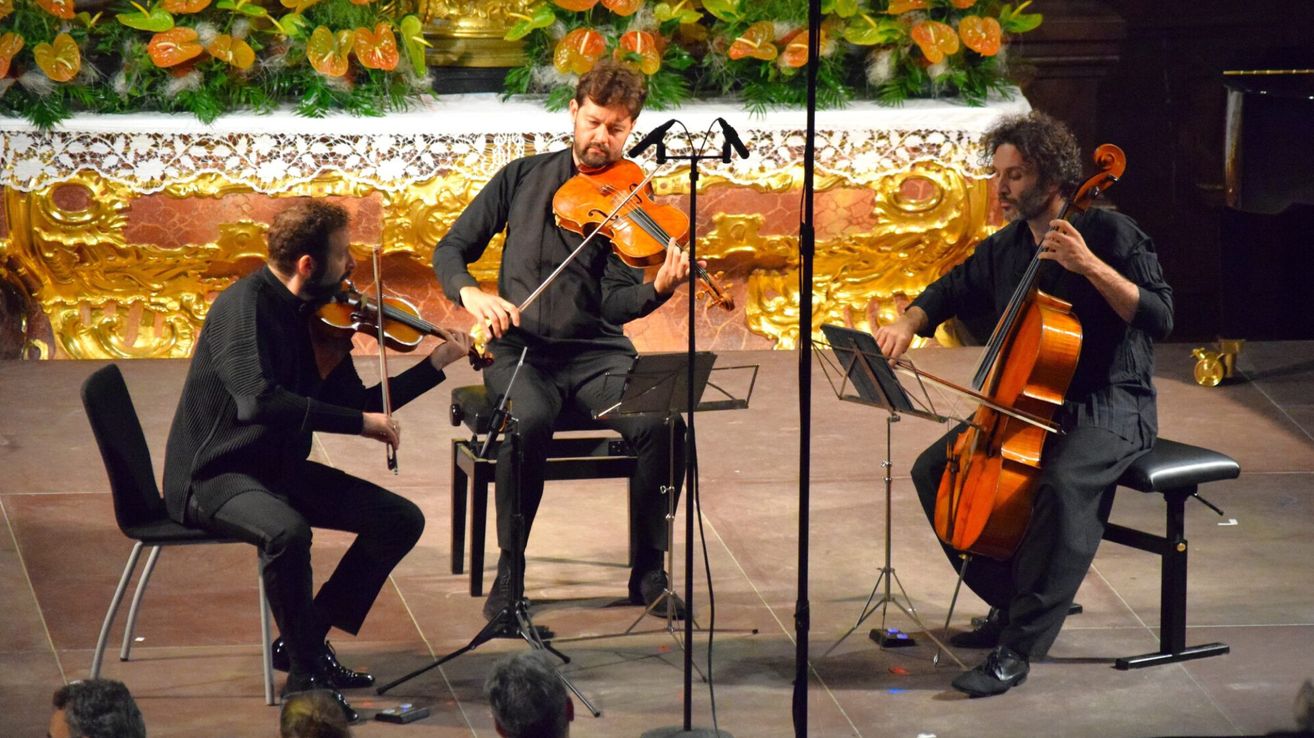 Drei Musiker sind auf der Bühne und spielen mit Streichinstrumenten. Der Mann in der Mitte spielt Geige, während der Mann auf der rechten Seite Cello spielt. Der Mann auf der linken Seite spielt Geige. Sie stehen vor einem Publikum.