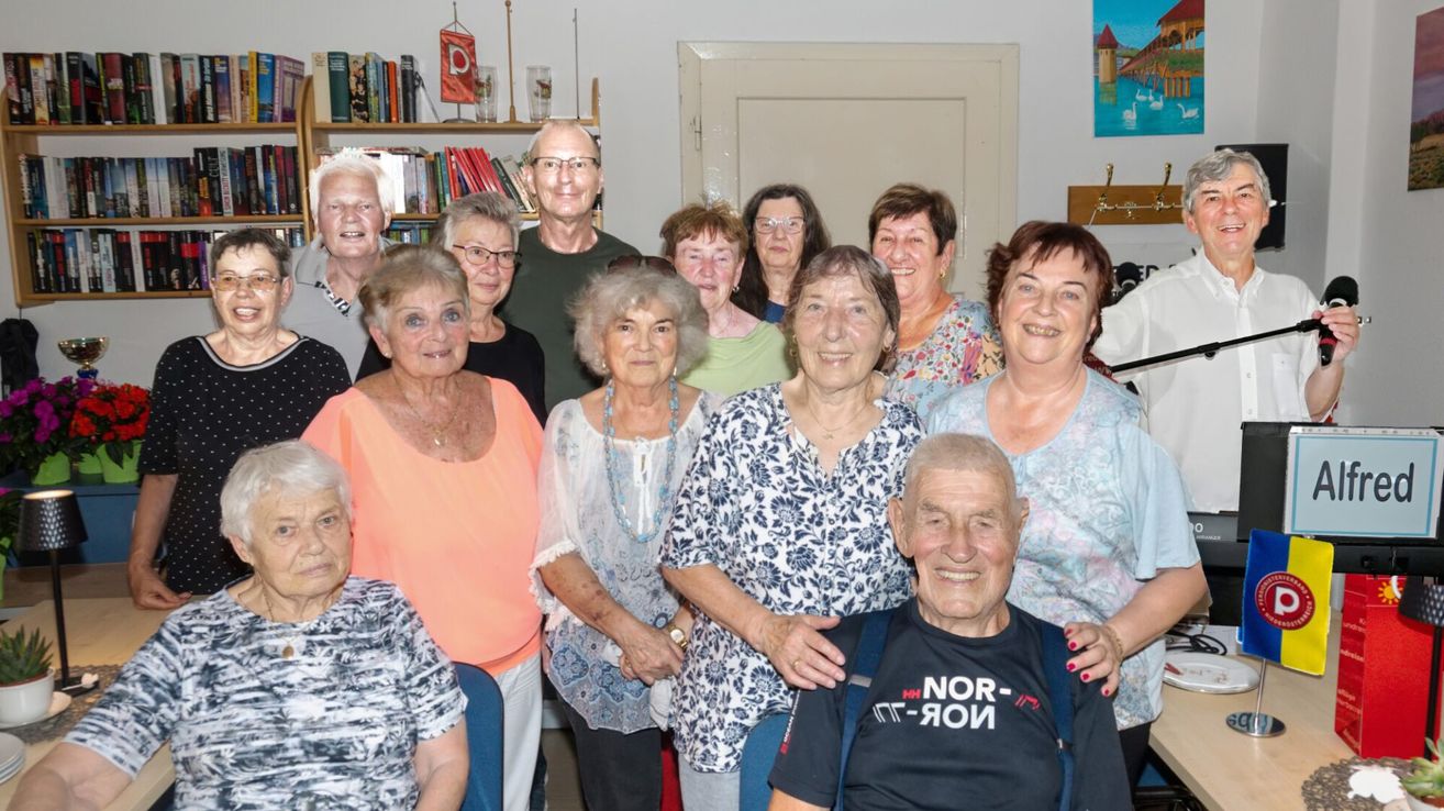 Eine Gruppe älterer Menschen, sowohl Männer als auch Frauen, versammelt sich in einem Raum, lächelt und posiert für ein Foto. Einige tragen Brillen, Halsketten und Armbänder. Ein Bücherregal mit Büchern und einer Flagge steht hinter ihnen.