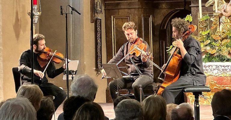 Drei Musiker spielen Violinen bei einer Aufführung vor einem Publikum, einer sitzt, zwei stehen. Sie haben Notenständer vor sich und ein Mikrofon auf einem Ständer in der Nähe.