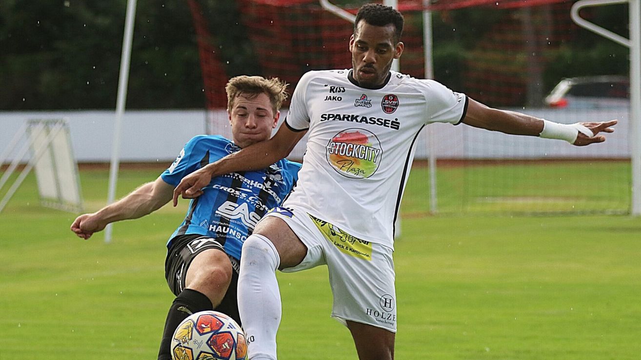 Zwei Fußballspieler sind auf dem Feld in Aktion. Einer in Weiß und der andere in Blau, sie kämpfen um den Ball.