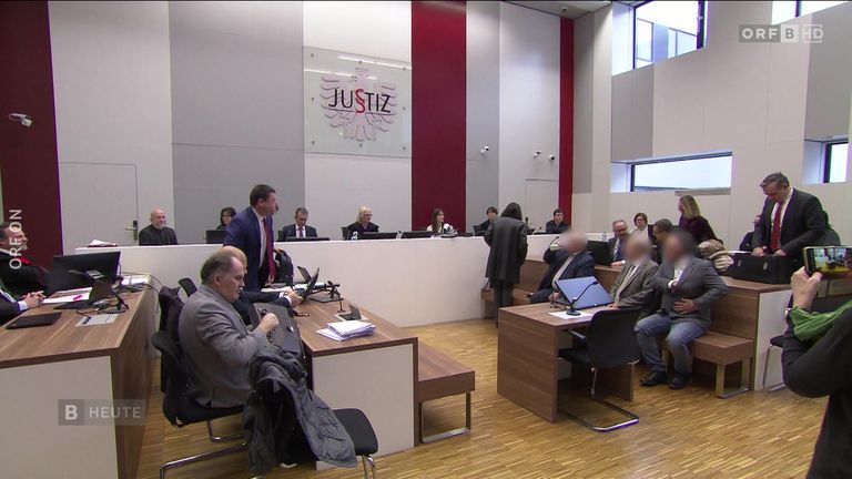 Eine Gruppe von Menschen sitzt in einem Gerichtssaal an Schreibtischen. Der Raum hat ein modernes Design mit Holzböden. Eine Glaswand zeigt das Wort 'Justiz' mit einem Adlerlogo.