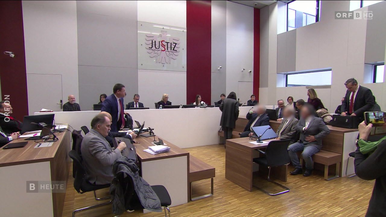 Eine Gruppe von Menschen sitzt in einem Gerichtssaal an Schreibtischen. Der Raum hat ein modernes Design mit Holzböden. Eine Glaswand zeigt das Wort 'Justiz' mit einem Adlerlogo.