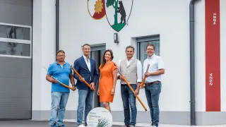 Fünf Personen posieren für ein Foto vor einem Gebäude. Sie halten alle Stöcke. Die Frau in der Mitte trägt ein oranges Kleid. Hinter ihnen befindet sich ein Logo an der Wand.