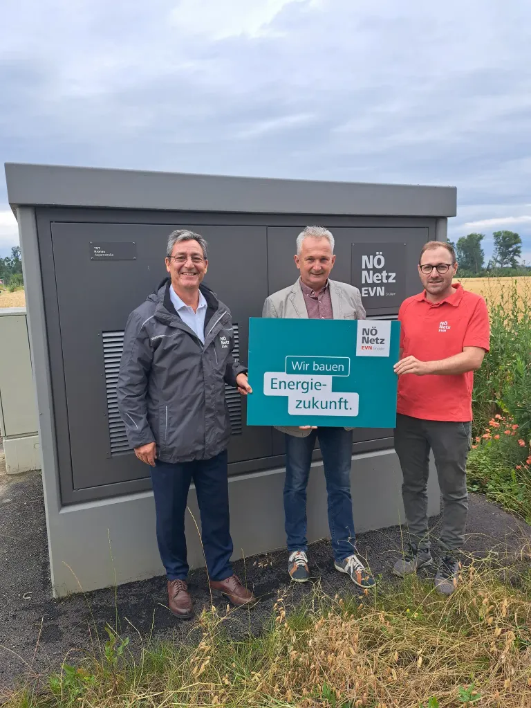 Drei Männer stehen vor einem grauen Stromverteilerkasten. Sie halten ein grünes Schild mit den Worten Wir bauen Energie-zukunft.