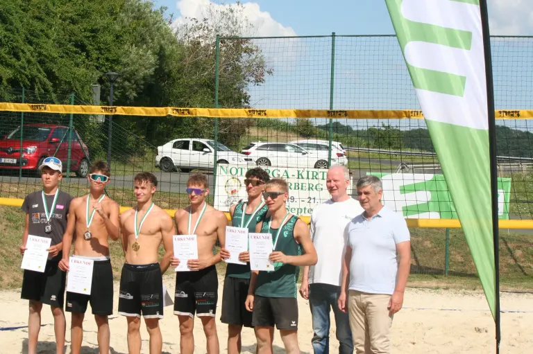 Fünf junge Männer in Sportuniformen und Medaillen posieren für ein Foto am Strand. Hinter ihnen steht ein Volleyballnetz und zwei Männer in Freizeitkleidung. Im Hintergrund sind geparkte Autos und Bäume zu sehen.