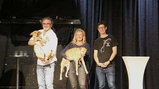 Drei Personen stehen lächelnd auf einer Bühne mit zwei Hunden. Der Mann auf der linken Seite hält einen Hund, die Frau in der Mitte hält einen Hund und der Mann auf der rechten Seite trägt eine Brille und ein schwarzes T-Shirt.
