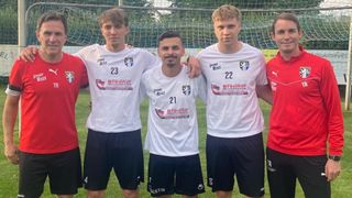 Vier Fußballspieler stehen zusammen auf einem Feld. Zwei Spieler haben ihre Arme umeinander. Ihre weißen Trikots tragen die Nummern 23, 21 und 22. Ein verschwommener Hintergrund zeigt ein Netz und Bäume.