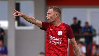 Ein Fußballspieler in einem roten Trikot mit Power Tech und Proyecto-Sponsoren gestikuliert mit seinem Arm.
