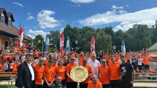 Eine Gruppe von Menschen in orangefarbenen Trikots feiert mit einer Trophäe auf einem Feld, während eine Menge hinter ihnen jubelt.