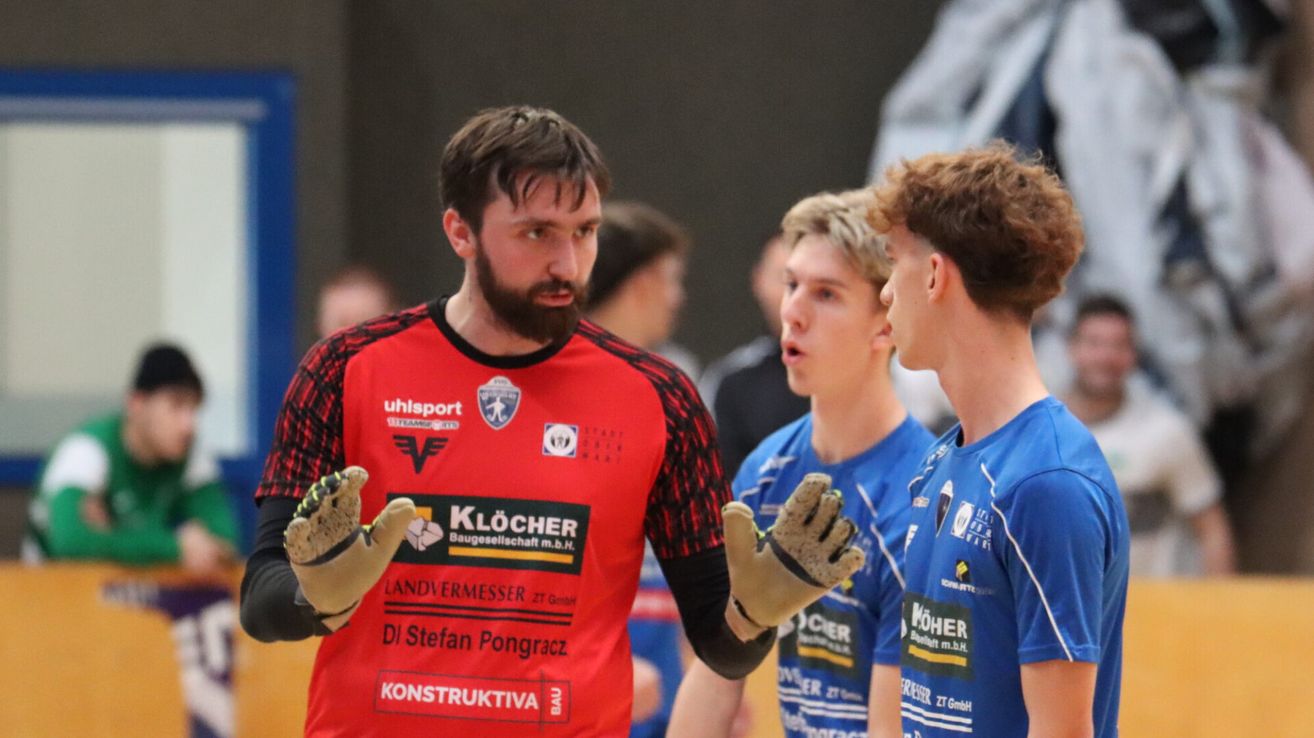 Eine Gruppe von Männern in Sportuniformen, einschließlich eines Mannes in Rot und Handschuhen, schauen sich gegenseitig an. Der unscharfe Hintergrund zeigt andere Personen und ein undeutliches Objekt.
