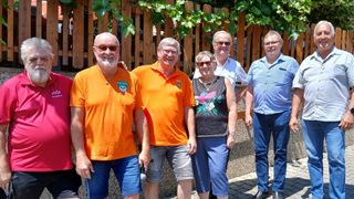 Fünf Personen stehen vor einem Holzzaun. Drei Männer tragen orangefarbene Hemden; einer hat ein Logo. Eine Frau in einem Blumenhemd und ein Mann in einem weißen Hemd stehen ebenfalls da.