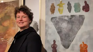 Eine Frau steht neben einer Wand mit zwei gerahmten Gemälden. Sie lächelt und trägt eine schwarze Jacke. Das Gemälde rechts hat einen großen grauen Felsen, mit kleineren farbigen Figuren darunter. Das Gemälde links hat ein abstraktes Design.