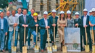 Gruppe von Personen in Schutzhelmen, die an einer Baustelle für ein Foto posieren, Schaufeln in der Hand, mit im Hintergrund fortlaufendem Bau.