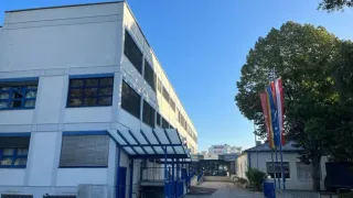 Ein großes weißes Gebäude mit blauen Verzierungen und mehreren Fenstern steht an einem sonnigen Tag. Ein Weg führt zum Eingang, in der Nähe stehen Fahnen an Stangen.