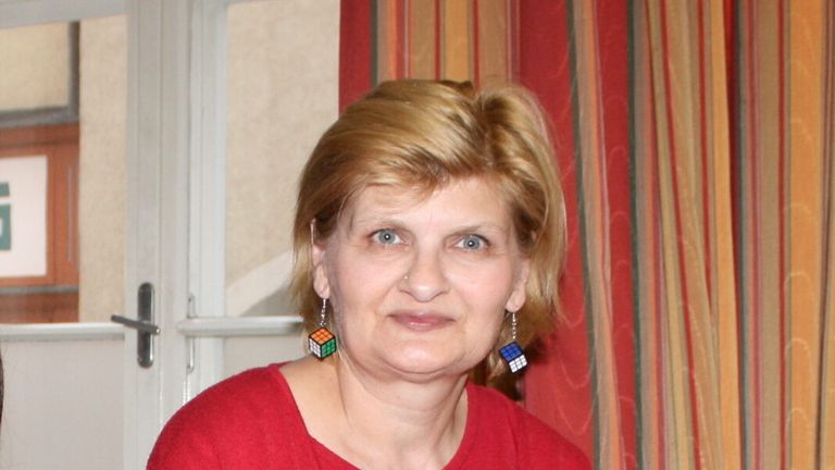 Eine Frau mit blondem Haar, die ein rotes Shirt und bunte Würfelohrringe trägt, posiert für ein Foto vor einem gestreiften Vorhang.