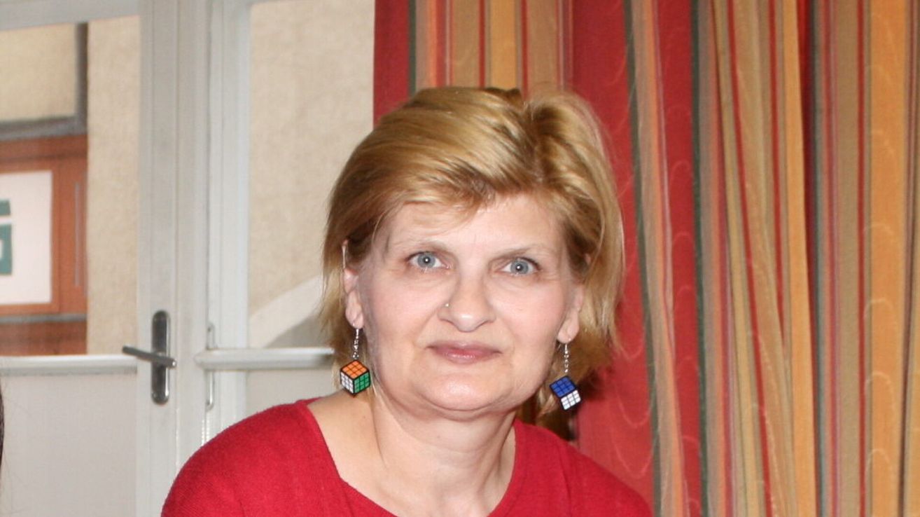 Eine Frau mit blondem Haar, die ein rotes Shirt und bunte Würfelohrringe trägt, posiert für ein Foto vor einem gestreiften Vorhang.