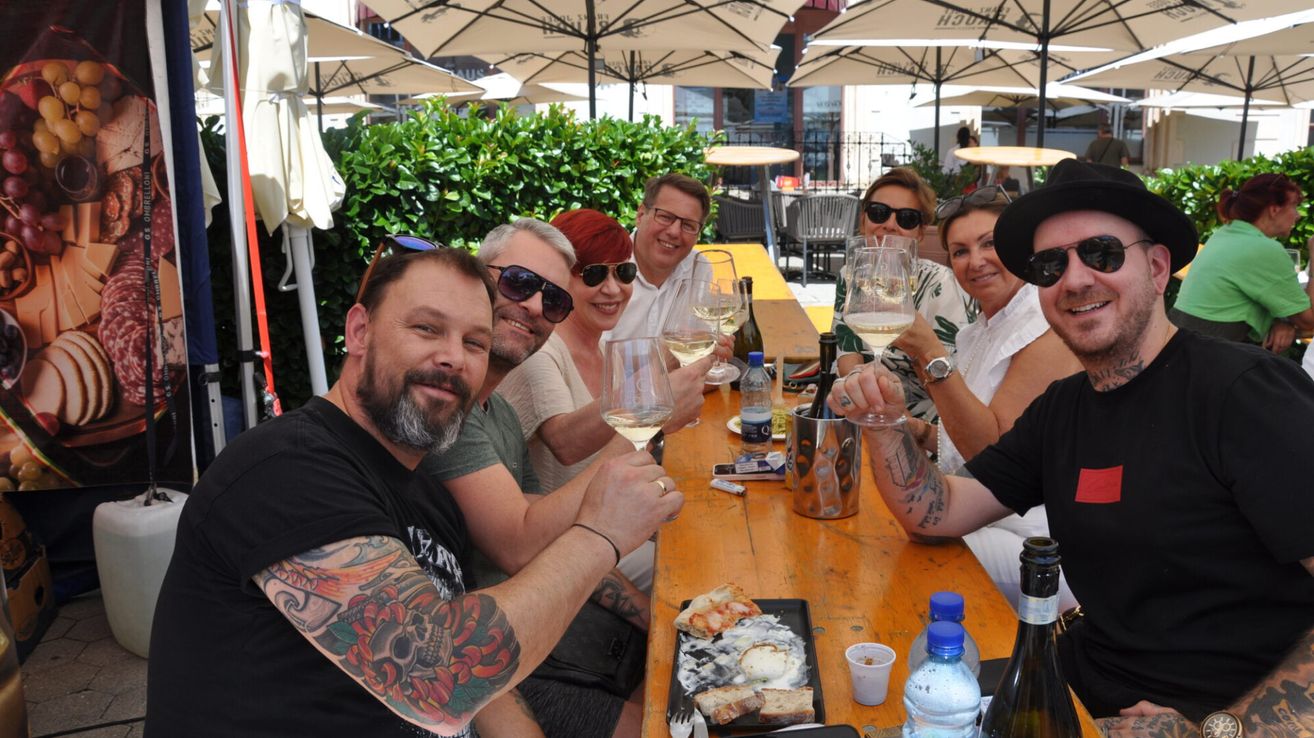 Eine Gruppe von Menschen, einige mit Tattoos, sitzt unter Schirmen um einen Tisch herum, hält Weingläser in der Hand, lächelt und posiert für ein Foto.