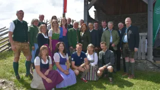 Eine Gruppe von Menschen in traditioneller bayerischer Kleidung posiert für ein Foto vor einem Holzgebäude. Die Gruppe besteht aus Männern und Frauen, von denen einige auf dem Gras sitzen.