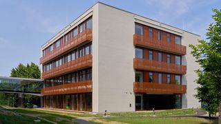 Ein modernes Gebäude mit beigefarbenen Wänden, Glasfenstern und roten Metallbalkonen. Es ist von Gras und Bäumen umgeben.