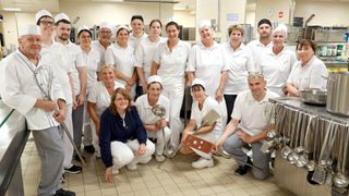 Eine Gruppe von Menschen in weißen Kochuniformen posiert für ein Foto in einer Küche, einige halten Küchenutensilien in den Händen.