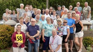 Eine Gruppe älterer Personen posiert für ein Foto auf Stufen. Einige tragen Brillen, Hüte und Uhren. Eine Person vorne hält ein gelbes Schild mit der Nummer 8.