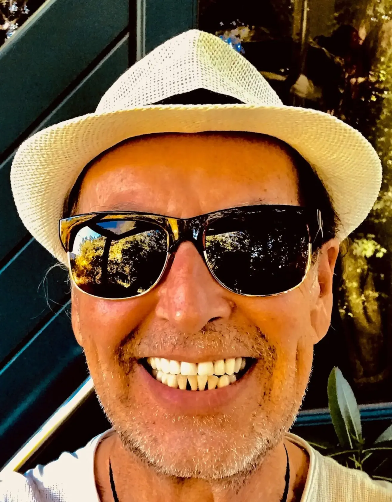 Ein lächelnder Mann mit Bart, der einen weißen Hut und Sonnenbrille trägt, reflektierte Bäume in den Gläsern.