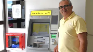 Ein älterer Mann mit Brille und gelbem Poloshirt steht neben einem weißen Geldautomaten mit einem gelben Schild, auf dem BankAutomatX steht.