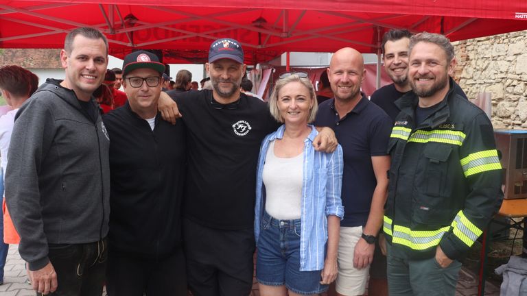 Eine Gruppe lächelnder Menschen unter einem roten Zelt. Die Frau in der Mitte trägt ein weißes Top und Jeansshorts. Ein Mann hinter ihr trägt die Uniform eines Feuerwehrmanns.