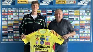 Zwei Männer posieren vor einer blauen Wand mit verschiedenen Logos. Der Mann auf der linken Seite hält ein gelbes Trikot mit den Worten Eggerglas Admiral. Der Mann auf der rechten Seite lächelt.