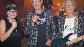 Drei Personen stehen in einem Raum zusammen und halten Mikrofone. Der Mann in der Mitte trägt eine karierte Jacke. Die Frau auf der linken Seite hält ein Mikrofon, und der Mann auf der rechten Seite spielt eine Gitarre.