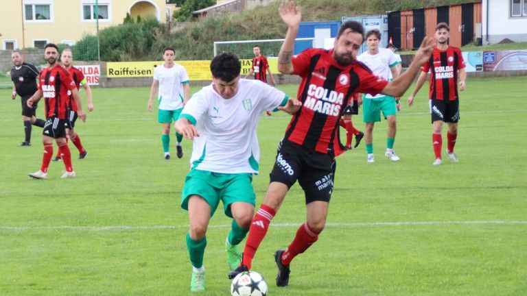 Fußballspieler in Aktion auf einem Feld, einer in Rot und Schwarz, der andere in Weiß und Grün. Der Spieler in Rot und Schwarz versucht, dem anderen Spieler den Ball zu stehlen.