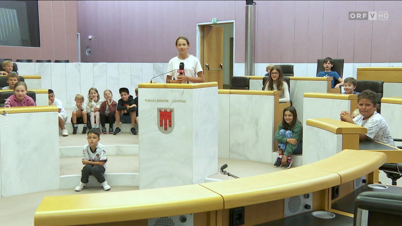 Ein junges Mädchen spricht an einem Rednerpult in einem Gesetzgebungssaal, mit Kindern um sie herum. Der Saal hat weiße Marmorwände und ein rotes Emblem. Ein Mikrofon ist vor ihr aufgestellt.