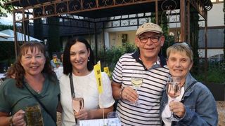 Vier Personen, zwei Frauen und zwei Männer, lächeln und posieren für ein Foto. Sie halten Gläser mit Wein und Bier. Hinter ihnen steht ein Pavillon mit einem Gebäude und Bäumen.